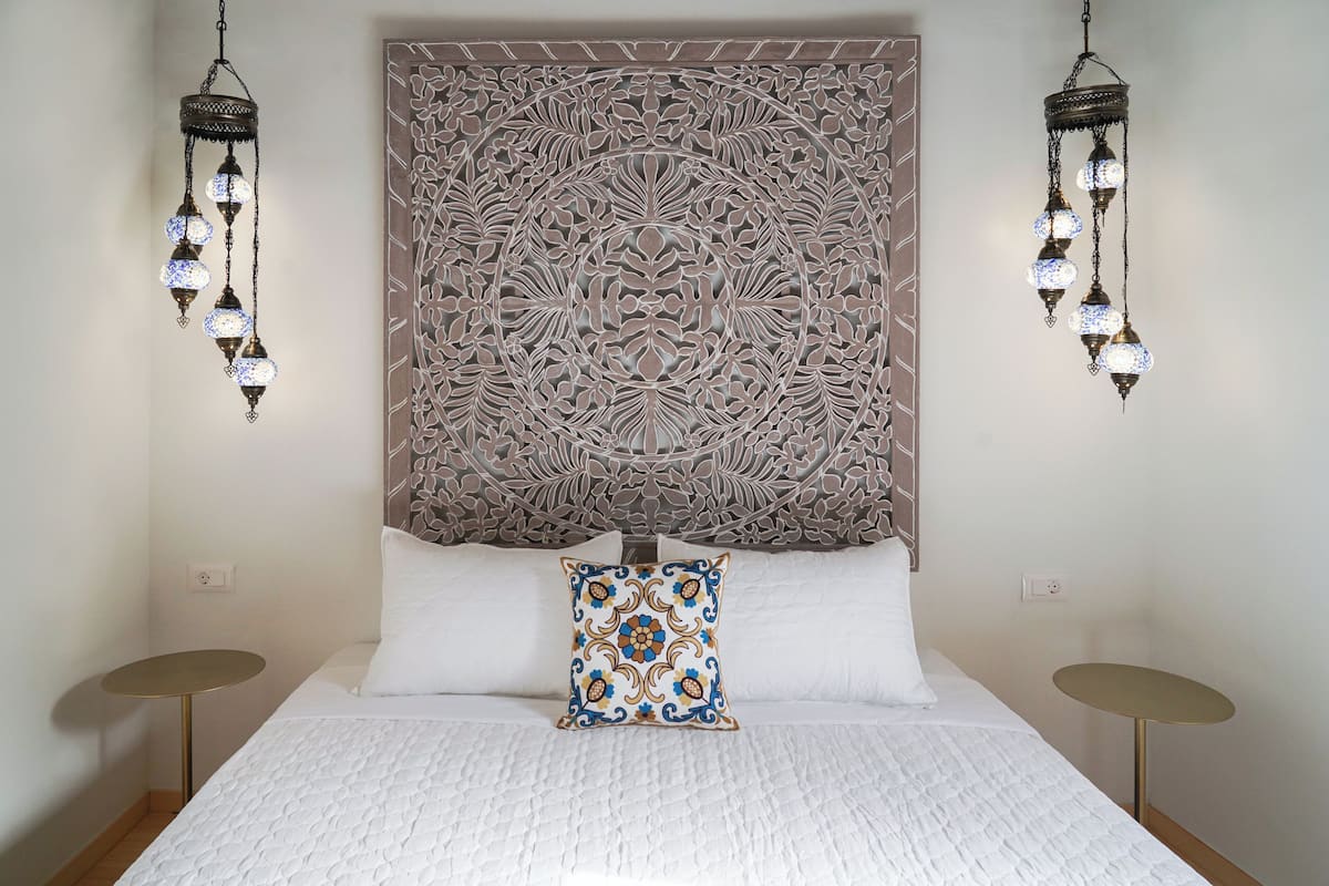 Cama bem arrumada com travesseiro decorativo, parede com desenhos elaborados e duas luminárias pendentes.