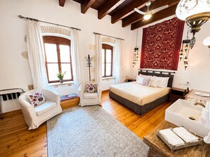 A bedroom with a bed, a chair, a bedside table, a lamp, a window, and a red tapestry. - Venetian Palace Chania (Chania)