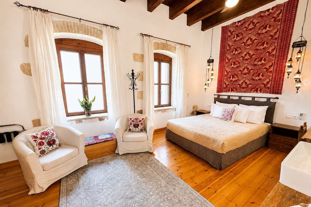 Quarto com uma cama grande, duas cadeiras, uma mesa pequena e uma janela ampla.