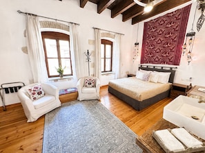 A room with a bed, two chairs, a small table, a bathtub, and a large window.