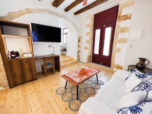 A living room with a wooden floor, a sofa with patterned cushions, a coffee table, a TV mounted on a wall, a red door, and a stone wall. - Venetian Palace Chania (Chania)