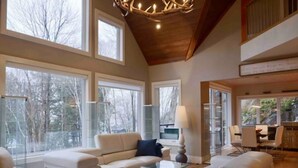 A spacious living room with large windows, a high ceiling, and a dining area with natural light. - The Rowanwood - Muskoka Cottage Retreat (Huntsville)