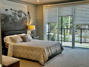 Ein Schlafzimmer mit einem großen Bett, einem Nachttisch, einer Lampe und Blick ins Freie durch große Fenster.