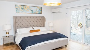 Quarto com cabeceira estofada, uma janela ampla com vista para árvores, mesa de cabeceira com abajures e um quadro na parede.