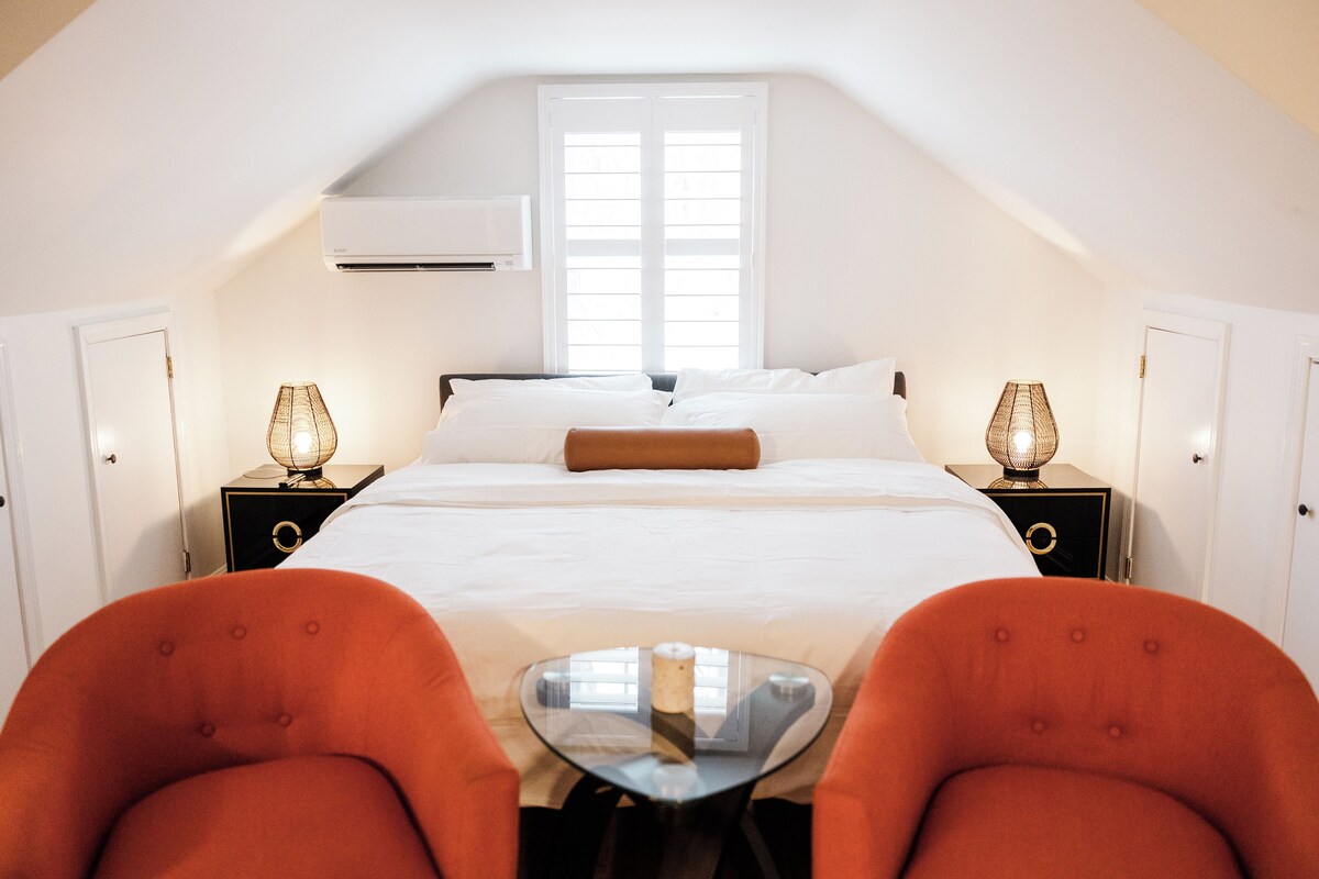 A bedroom with a large bed, two bedside tables with lamps, a small round table, and two orange chairs.