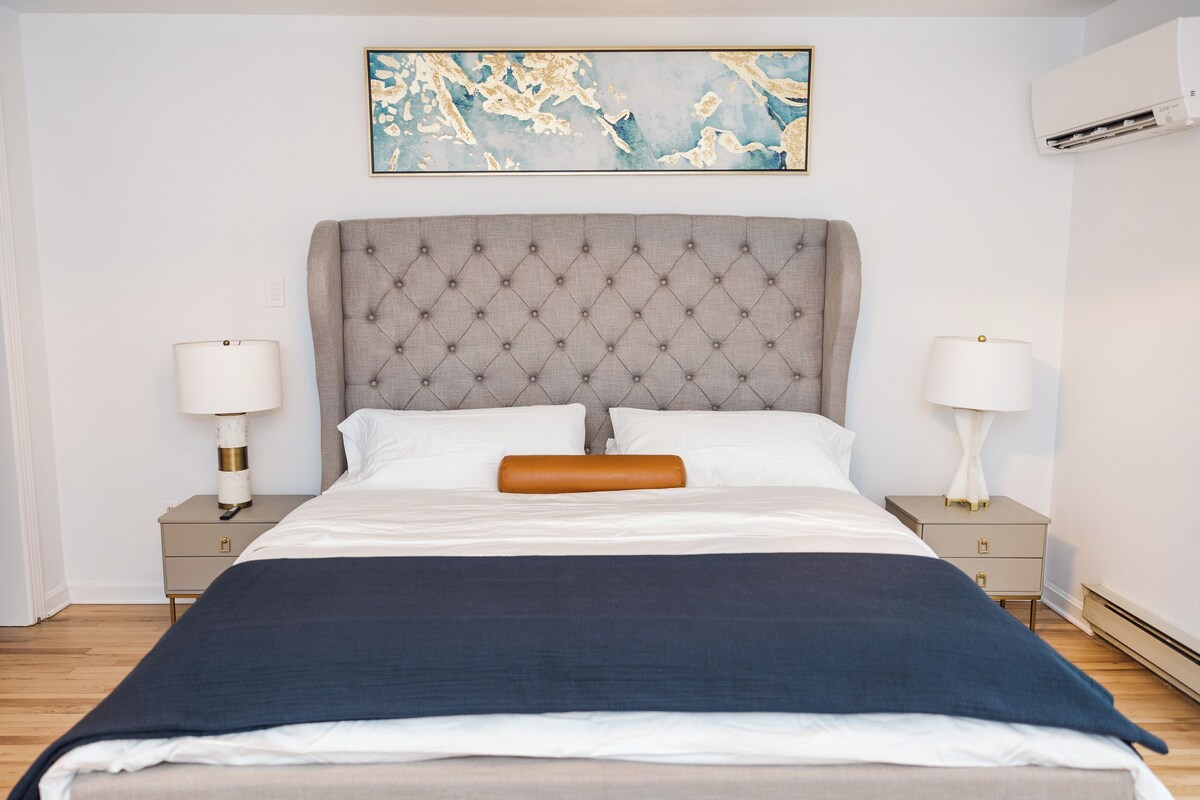 A neatly made bed with a tufted headboard, two bedside tables with lamps, and a wall-mounted air conditioner.