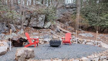 A campsite with two red Adirondack chairs, a fire pit, and a wooden bucket.
