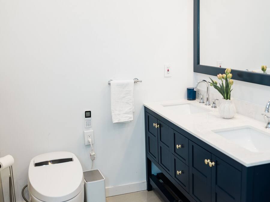 Een moderne badkamer met een wit toilet, een prullenbak, een wastafel met spiegel en een vaas met bloemen.
