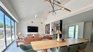 A spacious living area with a large dining table, comfortable seating, a flat-screen TV, and a view through large windows. - Austin Lux 4BR w/Wellness Center-Heated Pool/Sauna & Cold Plunge in Westlake (Austin)