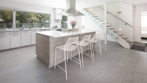 A modern kitchen with a central island, white bar stools, and a staircase leading to an upper level. - Austin Lux 4BR w/Wellness Center-Heated Pool/Sauna & Cold Plunge in Westlake (Austin)