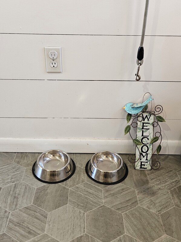 Una cocina con dos cuencos de acero inoxidable para mascotas sobre un suelo de baldosas hexagonales, un enchufe y un cartel que dice 'WELCOME' con una decoración de pájaro.