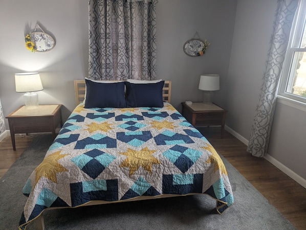 A bedroom with a bed, two bedside tables, lamps, a patterned quilt, and curtains.