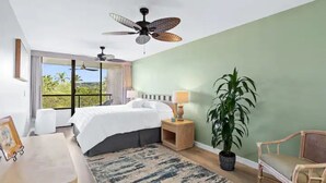 Een slaapkamer met een bed, nachtkastjes, een lamp, een stoel, een grote potplant en een plafondventilator.