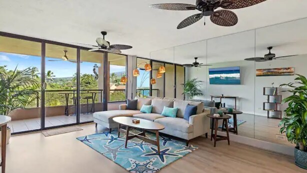Een ruime woonkamer met een grote bank, een salontafel en een balkon met uitzicht op palmbomen.