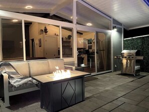 Une terrasse extérieure avec un canapé et un foyer, un barbecue, et une vue sur l’intérieur de la maison.