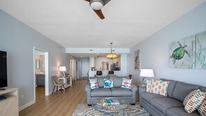A modern living room with a grey sofa, a glass coffee table, and a patterned rug. There is a dining area with a table and chairs, and a kitchen in the background.