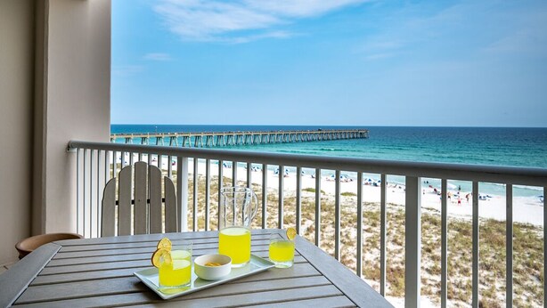 A balcony with a table set for drinks, overlooking a beach and a pier.