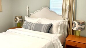Una cama bien hecha con sábanas blancas, una mesita de noche de madera con una lámpara y una ventana con cortinas.