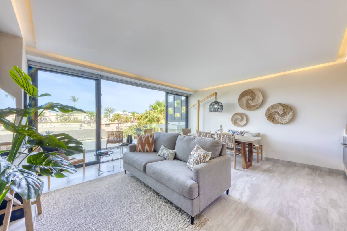 A modern living room with a grey sofa, a coffee table, and a dining area with a table and chairs. There are large windows offering a view of palm trees and buildings.
