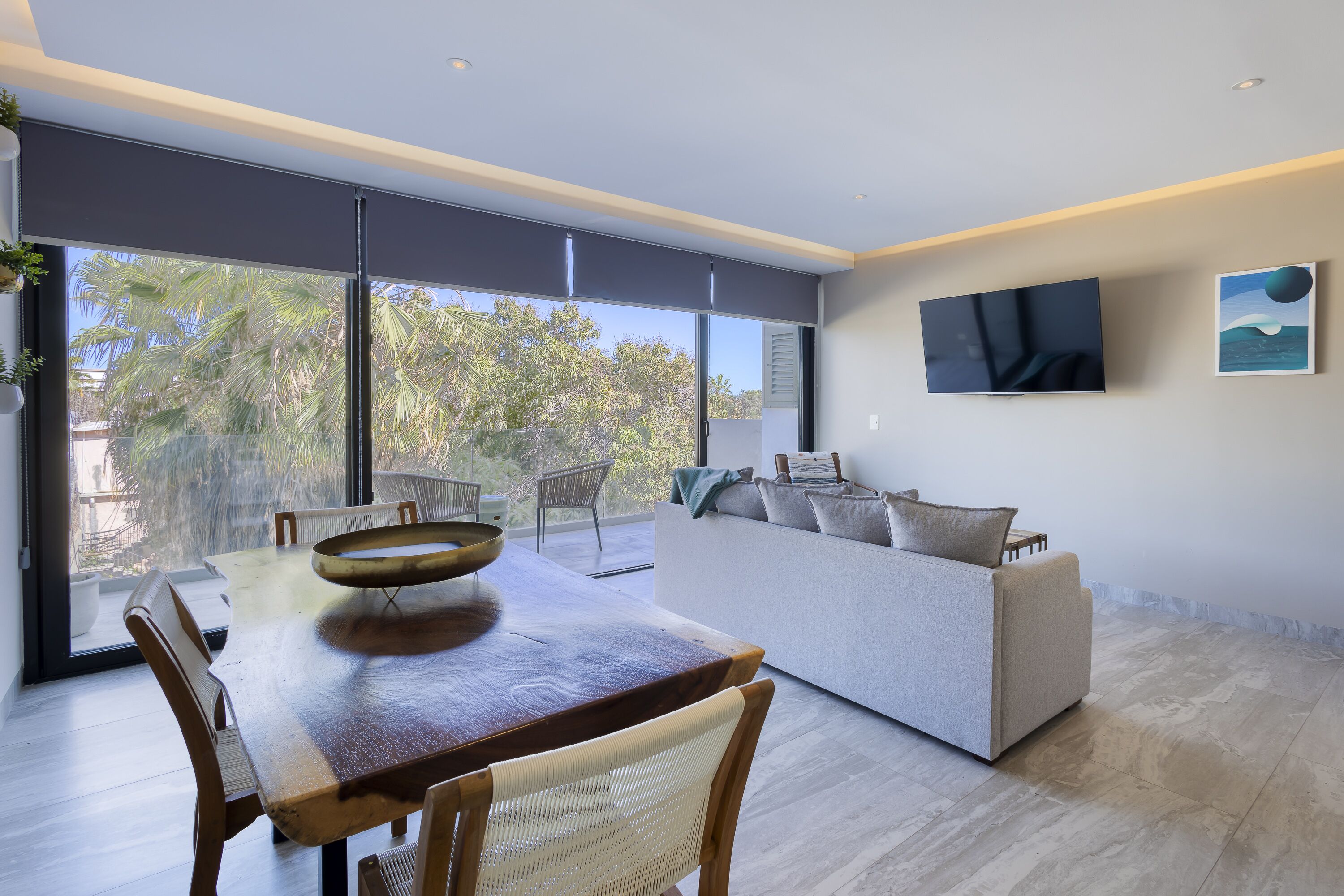 A modern living room with a dining area, large windows, a flat-screen TV, and a painting on the wall.