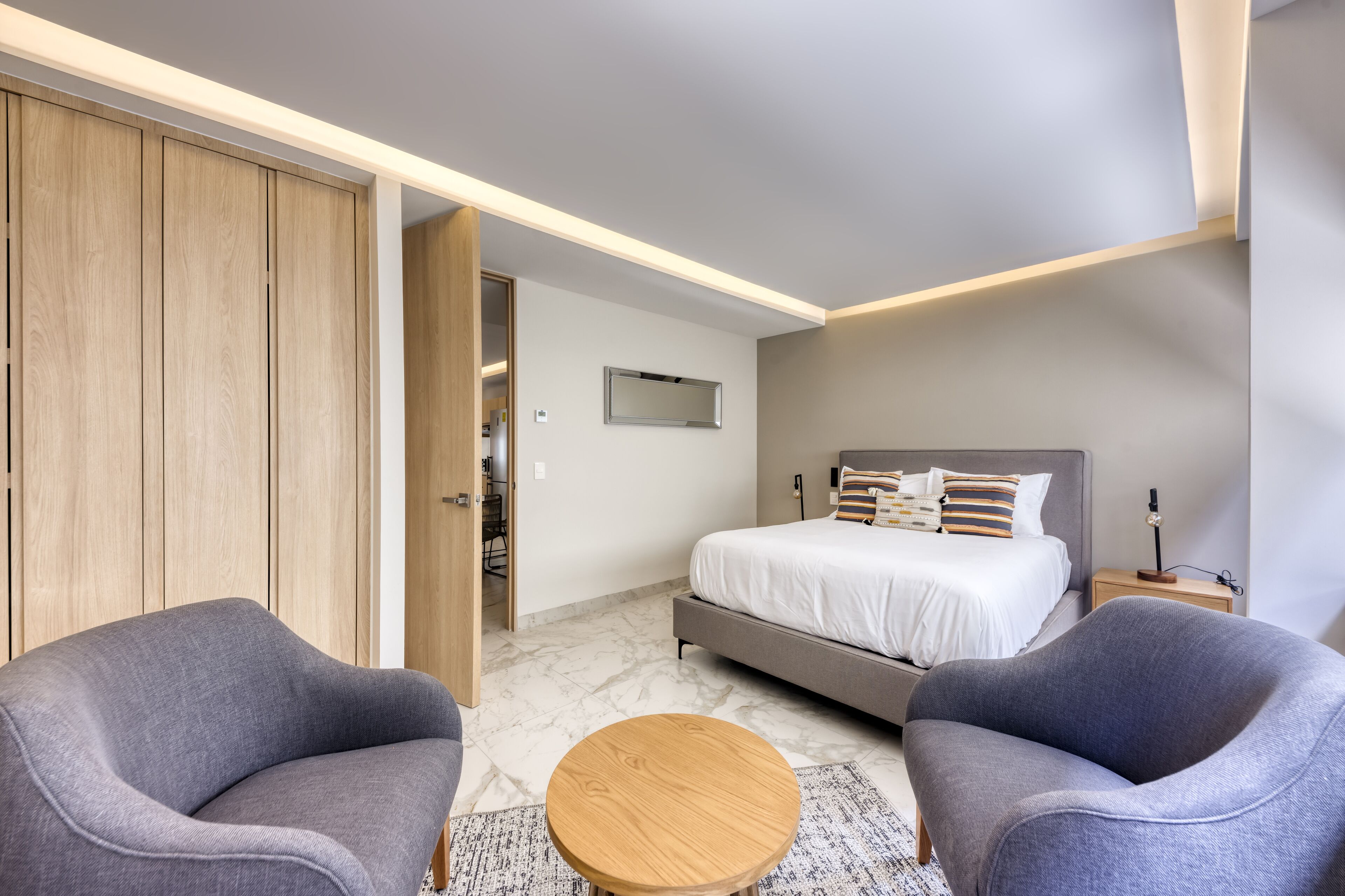 Un dormitorio moderno con una cama, dos sillones, una mesa redonda de madera y un armario.