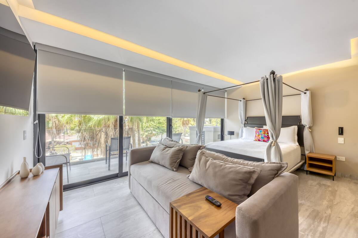 A modern hotel room with a large bed, a sofa, a small table, and a balcony with a view of greenery.