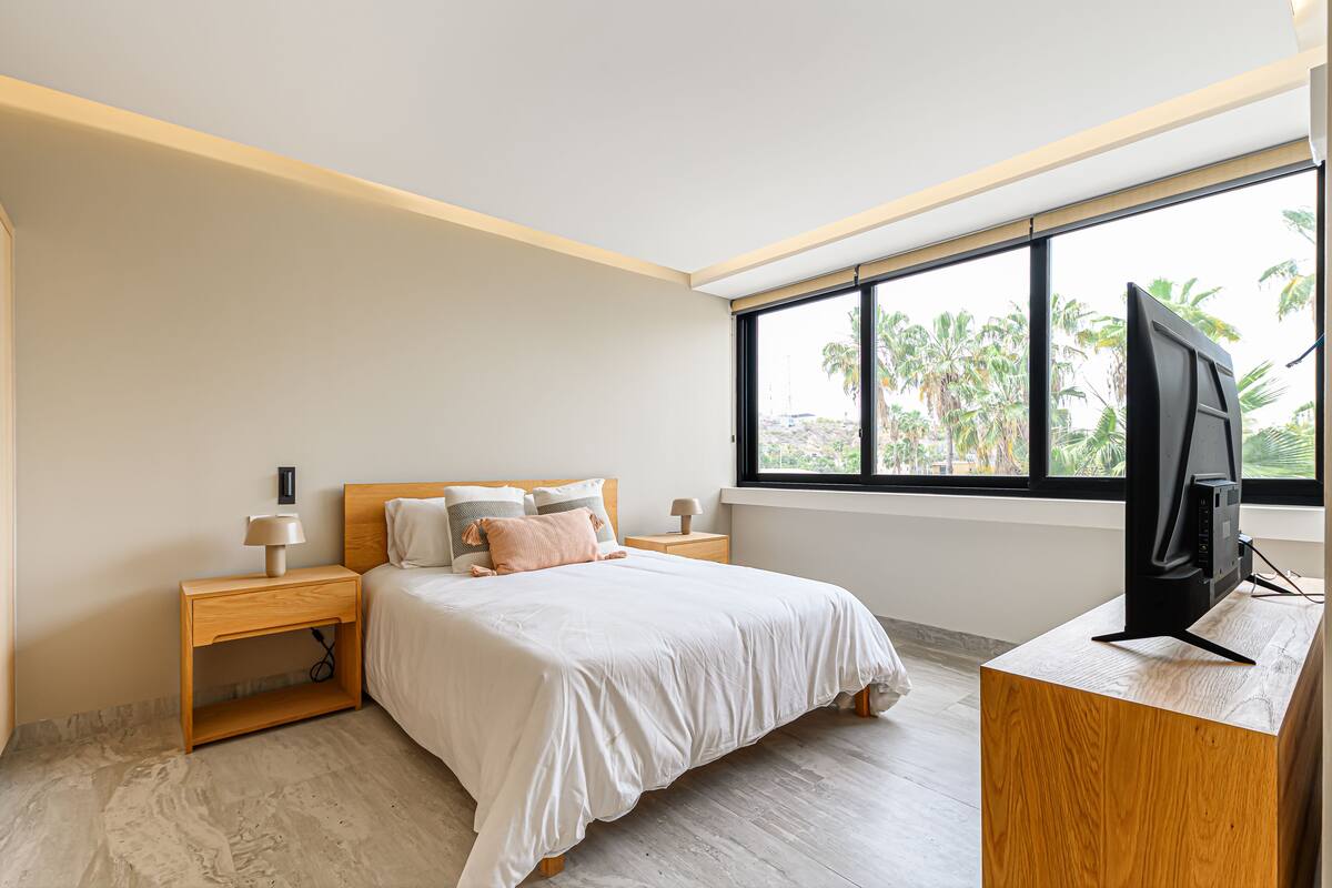 A modern bedroom with a large bed, wooden nightstands, a desk with a TV, and a window with a view of palm trees.