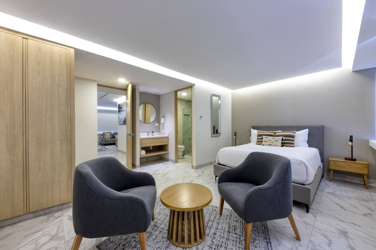 A modern hotel room with a large bed, two grey armchairs, a round wooden table, and a bathroom visible through an open door.