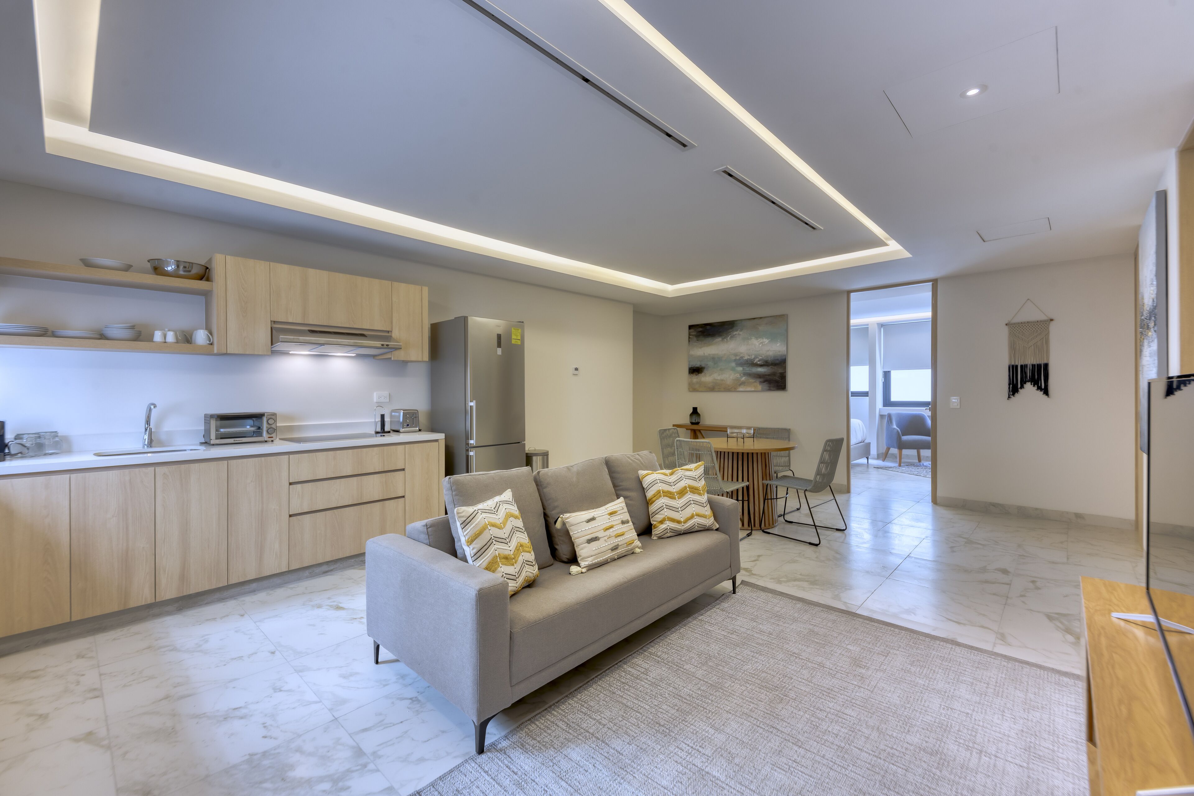 A modern kitchen with built-in appliances, a grey sofa with patterned cushions, and a dining area with a table and chairs.