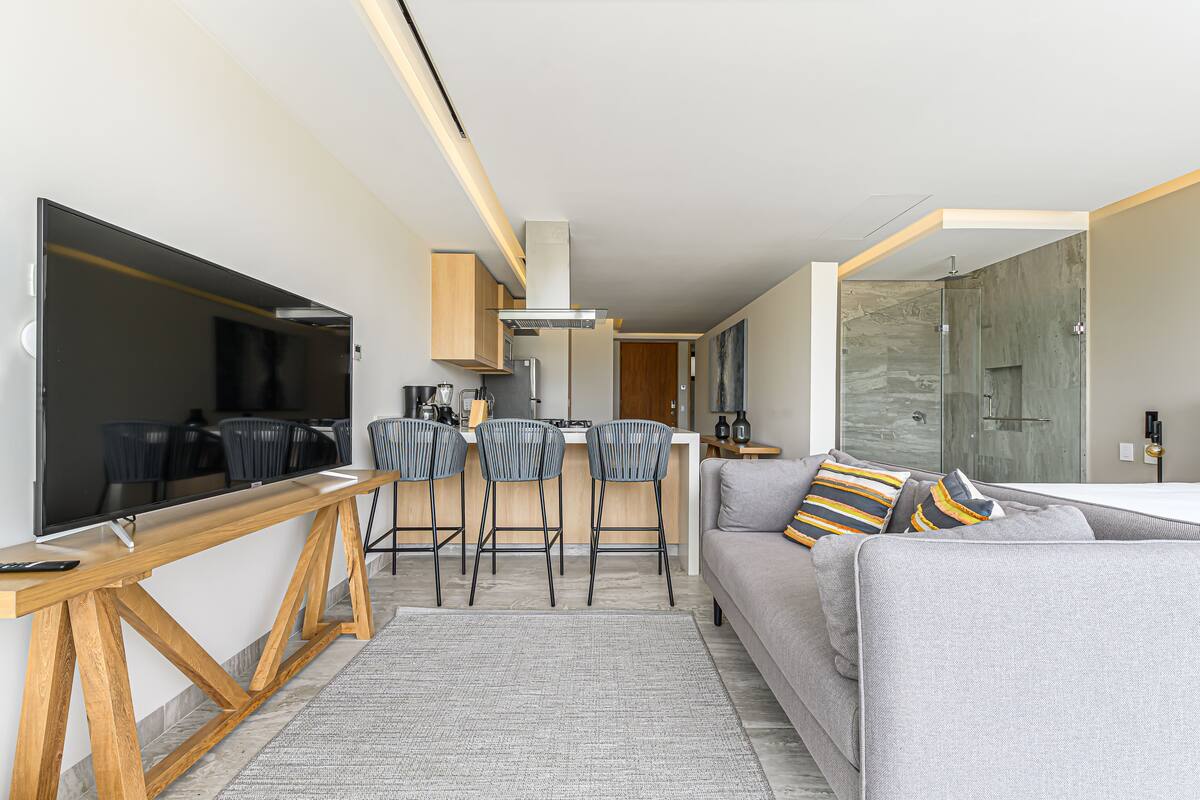 A modern living space with a flat-screen TV, a wooden dining table, a grey sofa with striped cushions, and a kitchen area with bar stools.