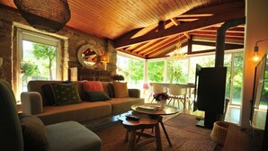 A cozy living room with a stone fireplace, wooden beams, a sofa, and a large window offering a view of greenery. - Casa da Luz Charming Cottage w/ Beautiful Garden (Vila Nova de Cerveira)