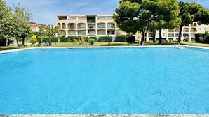 Una piscina con superficie de agua azul cristalina, rodeada por un césped cuidado y árboles. Al fondo, un edificio de varios pisos con balcones y fachada color crema.