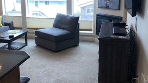 A living room with a gray sofa, a coffee table, and a fireplace. There are large windows with a view of buildings outside. - Our Happy Place Steps from Rockaway Beach (Rockaway Beach)