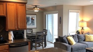 A modern kitchen with granite countertops, wooden cabinets, and a dishwasher. There is a dining area with a table and chairs, and a living room with a gray sofa and yellow pillows. - Our Happy Place Steps from Rockaway Beach (Rockaway Beach)