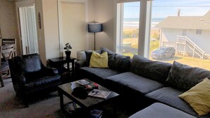 A living room with a sofa, armchair, and coffee table. There is a lamp and a plant on a side table. A window offers a view of a house and a car. - Our Happy Place Steps from Rockaway Beach (Rockaway Beach)