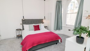 Un dormitorio con una cama tapizada en gris, ropa de cama blanca, una manta roja, un cabecero gris, dos mesitas de noche, una lámpara de pie, una ventana con cortinas verdes y una planta en maceta sobre un tocador blanco.