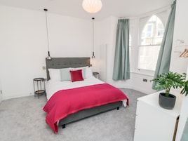 A bedroom with a grey upholstered bed, white bedding, a red throw blanket, a grey headboard, two bedside tables, a floor lamp, a window with green curtains, and a potted plant on a white dresser.
