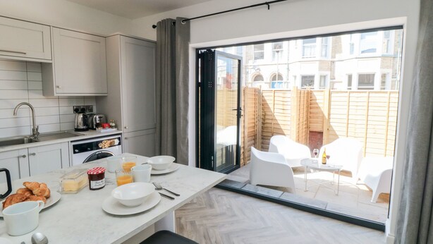 Una cocina moderna con armarios blancos, una zona de comedor con mesa dispuesta para el desayuno y un ventanal corredizo que da a un área de estar exterior.