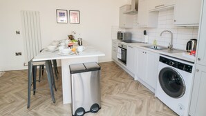 Cocina compacta con armarios blancos, horno empotrado, microondas y lavadora. Cuenta con una pequeña mesa de comedor con sillas y un cesto de basura.