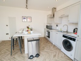 A compact kitchen with white cabinets, a built-in oven, a microwave, and a washing machine. There is a small dining table with chairs and a trash bin.
