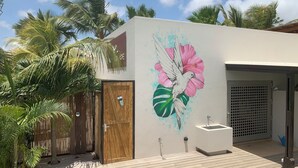 Un edificio moderno con una porta in legno, un muro bianco decorato con un murale di un uccello e fiori, un lavandino esterno e un patio in legno.