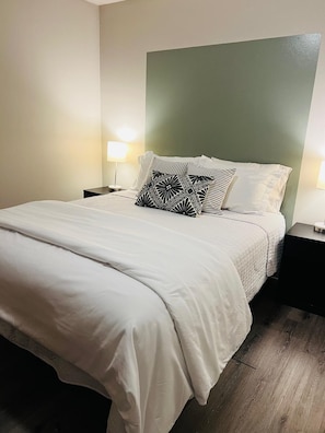 Un lit bien fait, avec une literie blanche et un coussin à motifs géométriques, flanqué de deux tables de chevet avec des lampes, dans une pièce dont un mur est peint dans une teinte verte.