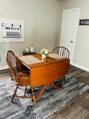 A dining room with a wooden table, four chairs, a vase with flowers, a wall-mounted air conditioner, and a door with a sign.