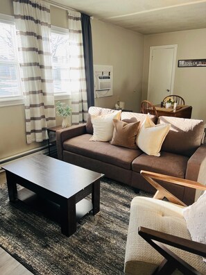 Ein Wohnzimmer mit Sofa, Couchtisch, Sessel, einem kleinen Tisch mit Vase und einer Wand-Klimaanlage.