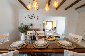 Uma área de refeições com uma mesa de madeira posta para seis pessoas, rodeada por cadeiras brancas. Há lâmpadas penduradas sobre a mesa, e uma planta é visível ao fundo.
