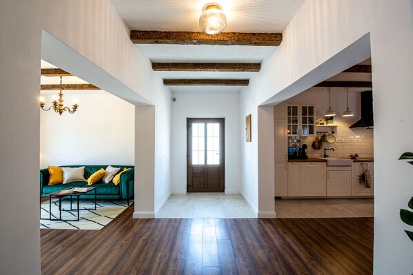 Um corredor amplo com piso de madeira, um sofá verde com almofadas amarelas, um candelabro e uma área de cozinha com armários brancos e uma coifa preta.