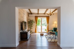 Um corredor com piso de madeira, parede de pedra, um vaso com planta, um espelho e vista para um quarto com uma poltrona verde e uma luminária.