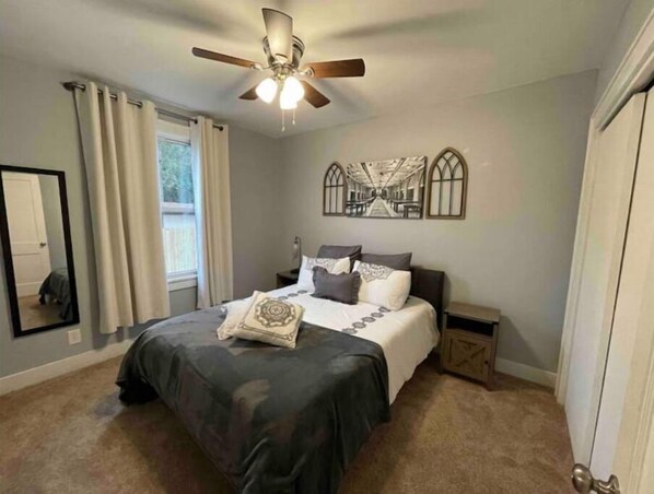 A bedroom with a bed, a ceiling fan, a nightstand, a mirror, and a window with curtains.