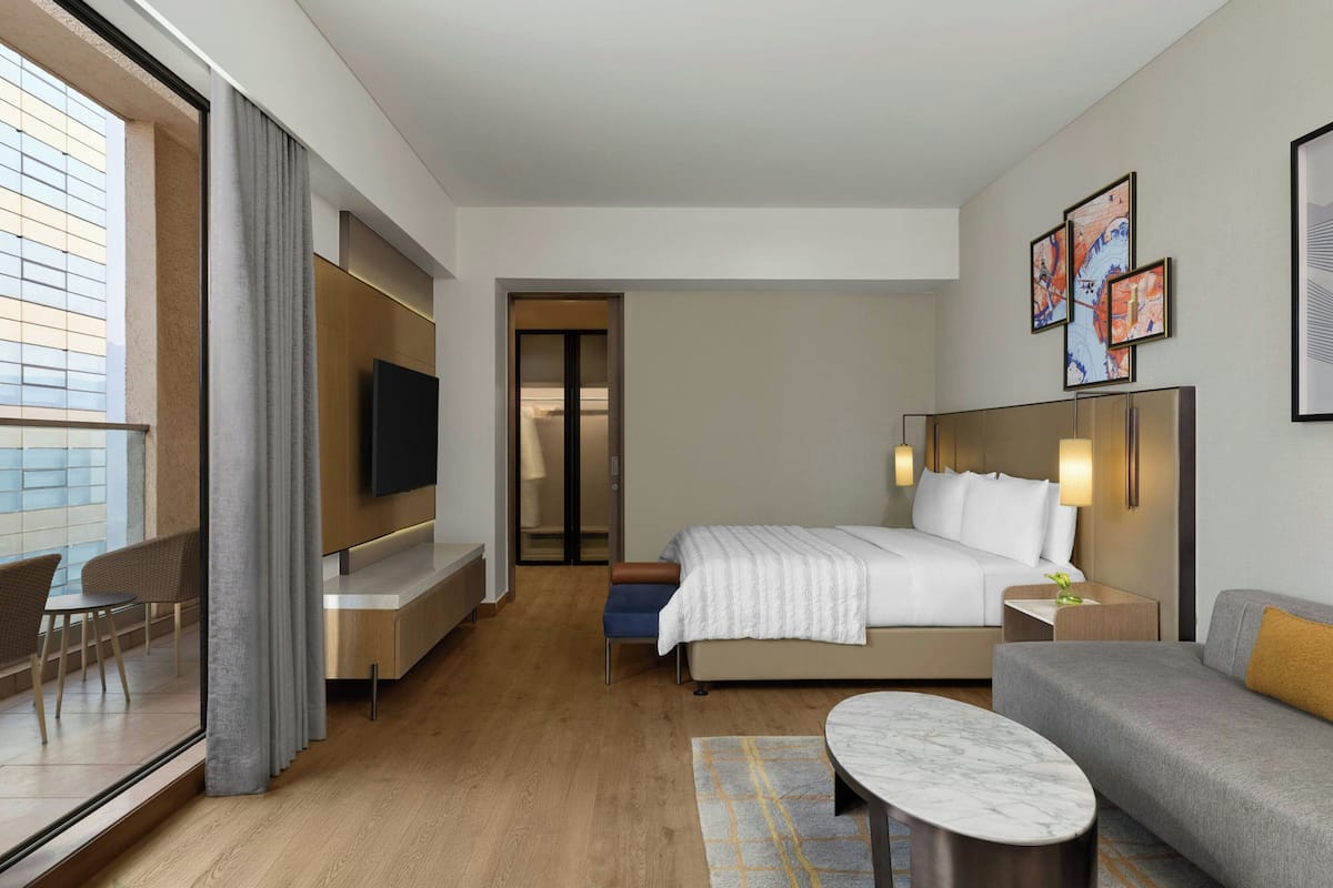 Een moderne hotelkamer met een bed, een televisie, een bank, een klein tafeltje en uitzicht op de stad door grote ramen.