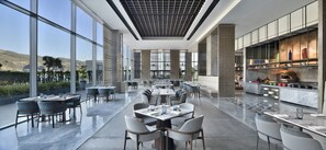 A modern restaurant with a long dining area, large windows, and a kitchen visible on the right. - Le Méridien Navi Mumbai (Navi Mumbai)